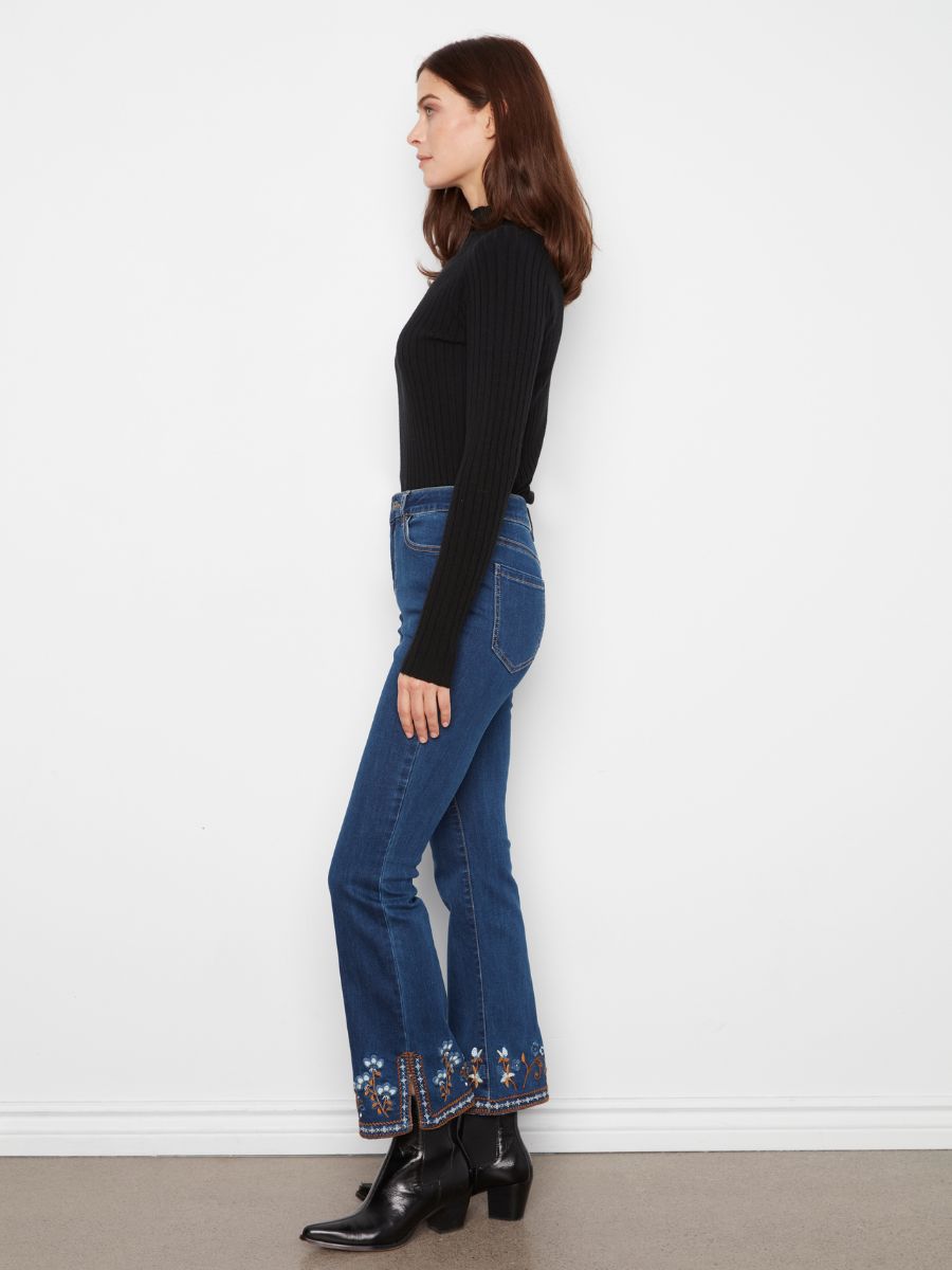 Side view of a figure wearing indigo denim jeans with floral embroidery at the hem paired with black heeled boots and a black ribbed top.