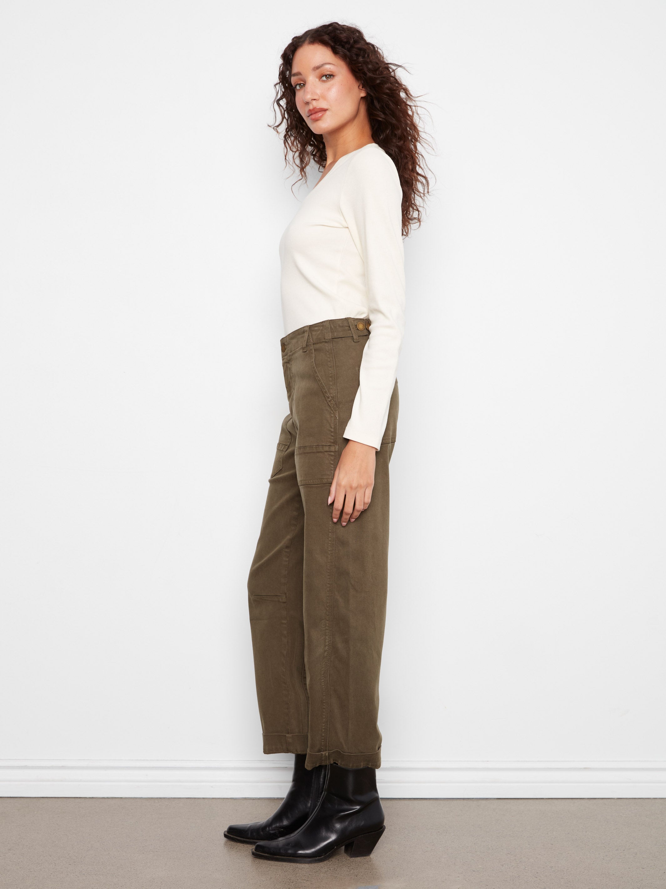 Side view of a figure dressed in a long-sleeve cream top, olive wide-leg pants, and black ankle boots against a white wall.