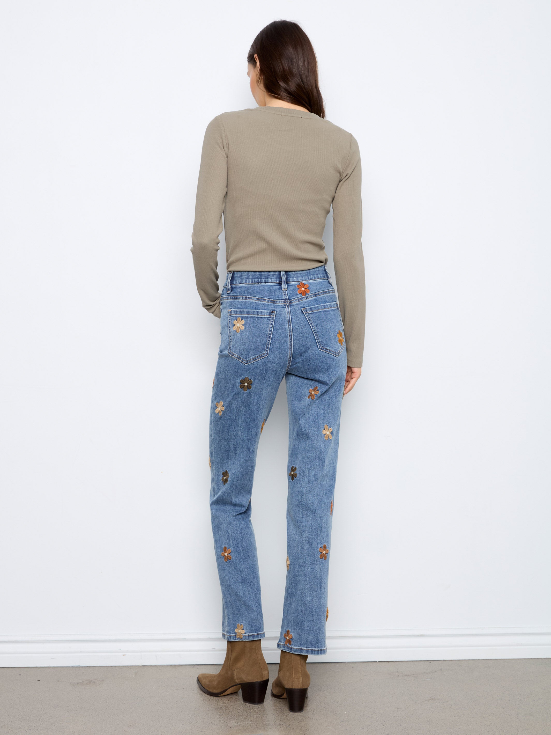 Back view of light blue denim jeans with multicolored floral embroidery paired with brown boots and a taupe long-sleeve top.