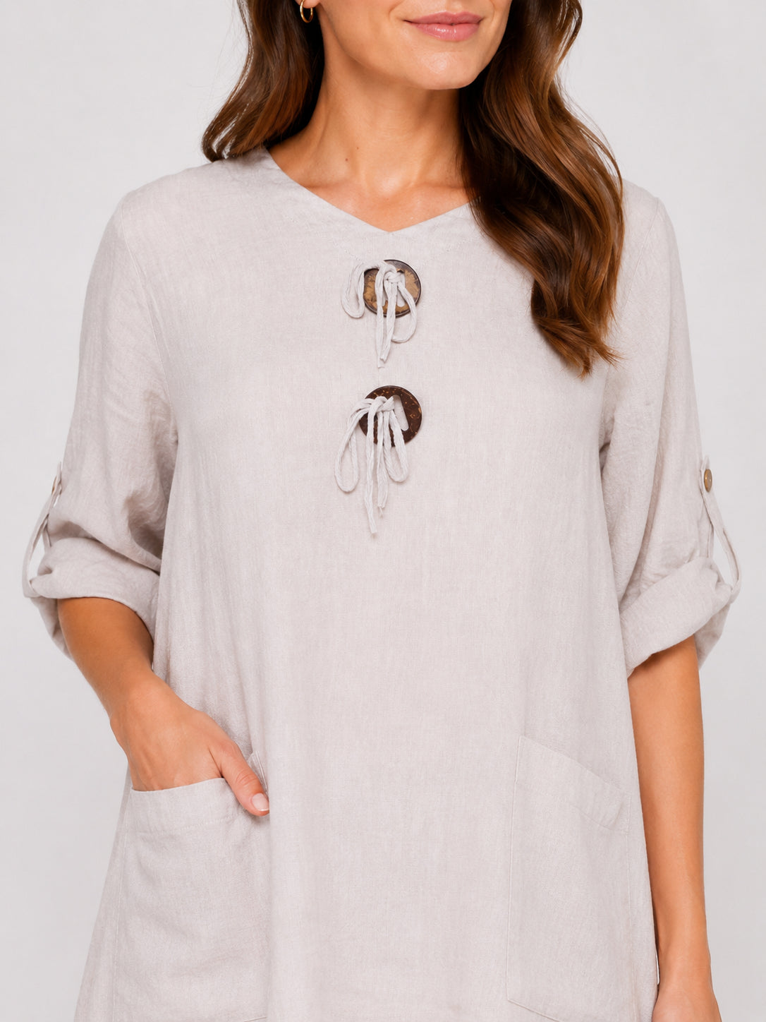 Beige linen long-sleeve top with rolled sleeves, front pockets, and two decorative circular tie details at the neckline.