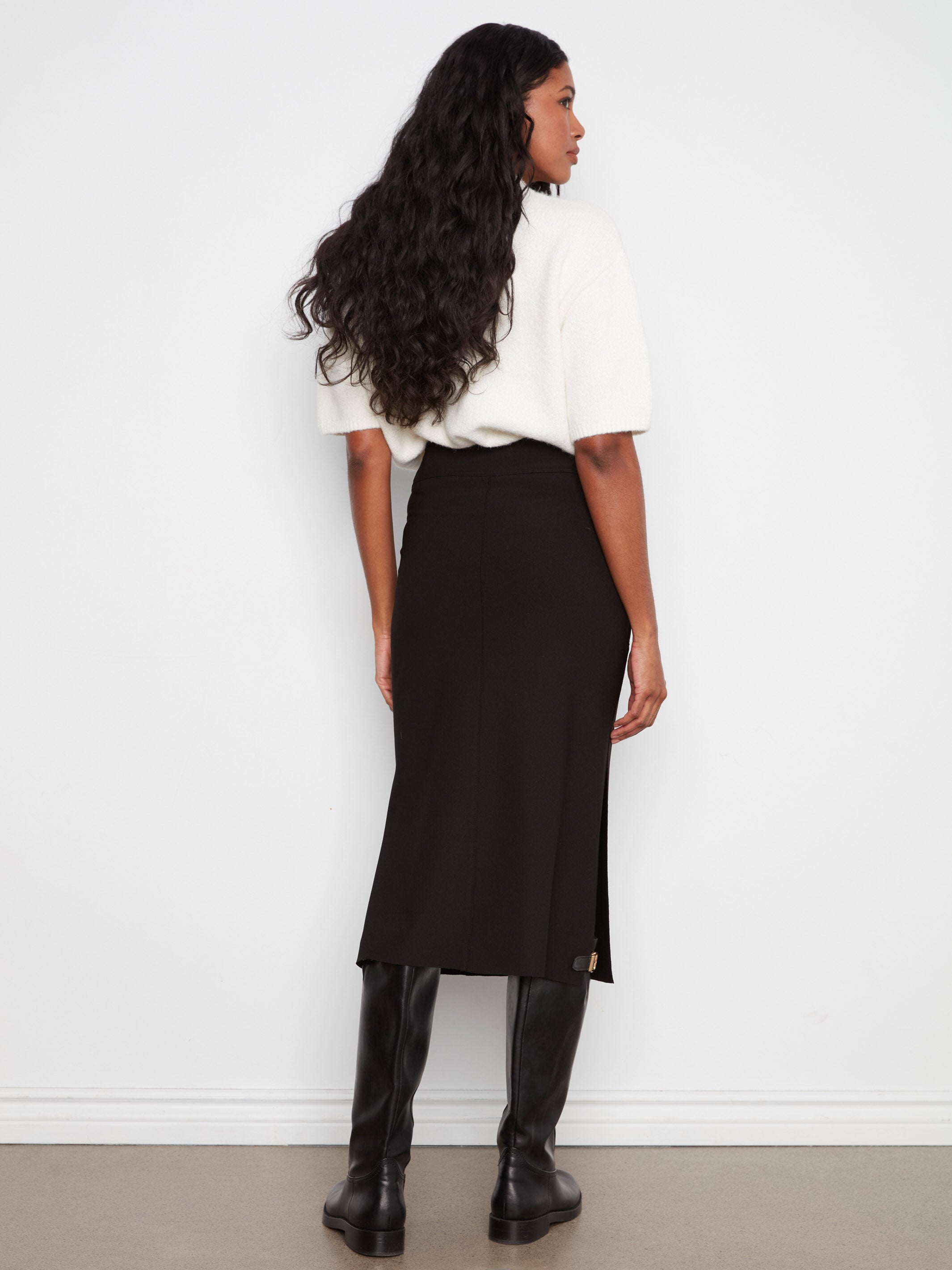 Back view of a figure in a white short-sleeve top, black midi skirt, and tall black boots against a plain wall.