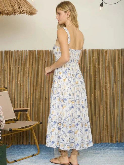 Sleeveless midi dress with blue and yellow tropical print and smocked back detail on model standing indoors