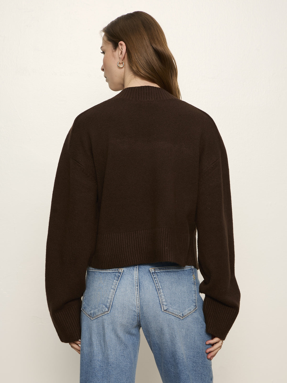Back view of a person wearing a dark brown knit sweater with ribbed cuffs and hem paired with light blue denim jeans.