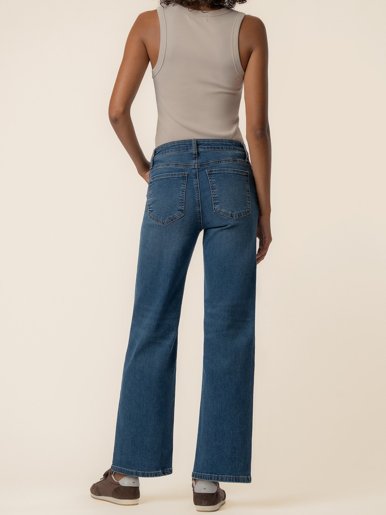 Rear view of a sleeveless beige top paired with high-waisted straight-leg blue denim jeans and brown sneakers.