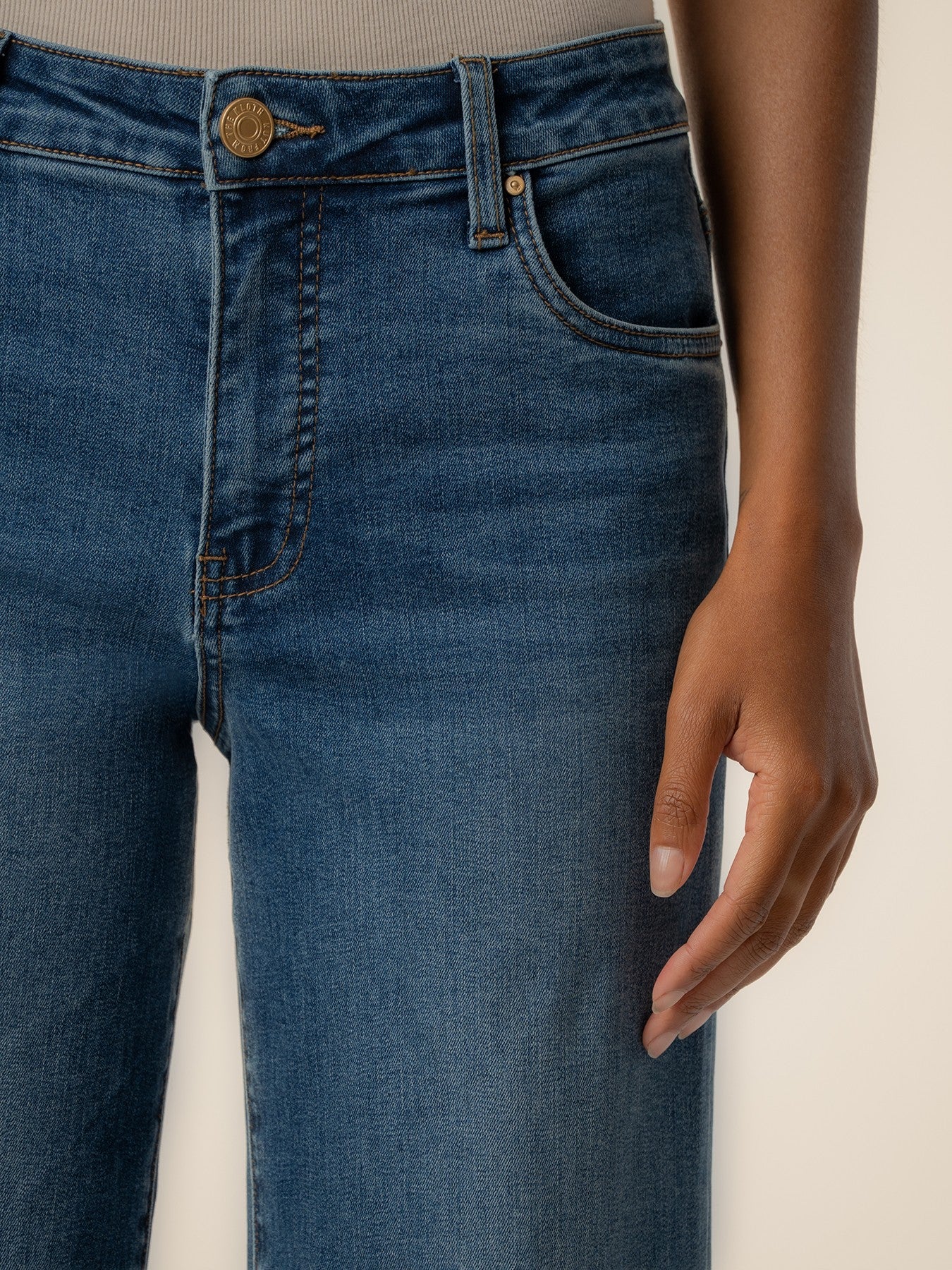Close-up of mid-blue denim jeans with detailed stitching and a brass button closure on a light background.