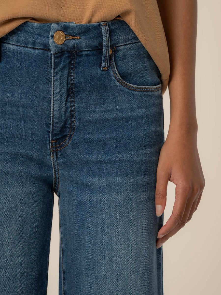 Close-up of blue denim Kut from the Kloth Meg 29” Wide Denim Jeans with a tucked beige top and hand resting at the side.