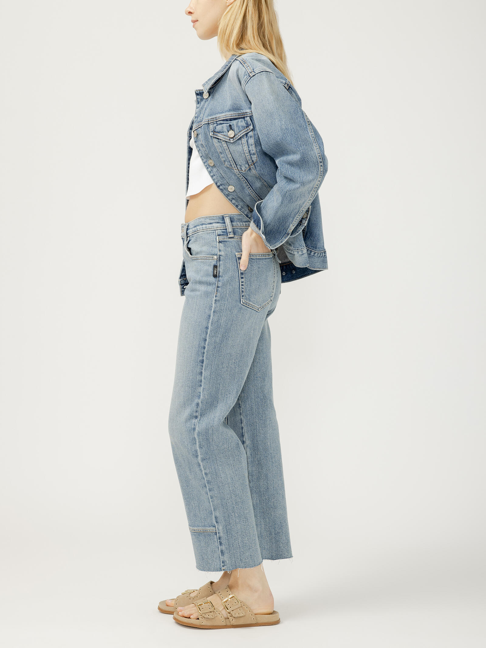 Side view of a light blue denim jacket and jeans outfit paired with beige buckle sandals against a plain background.