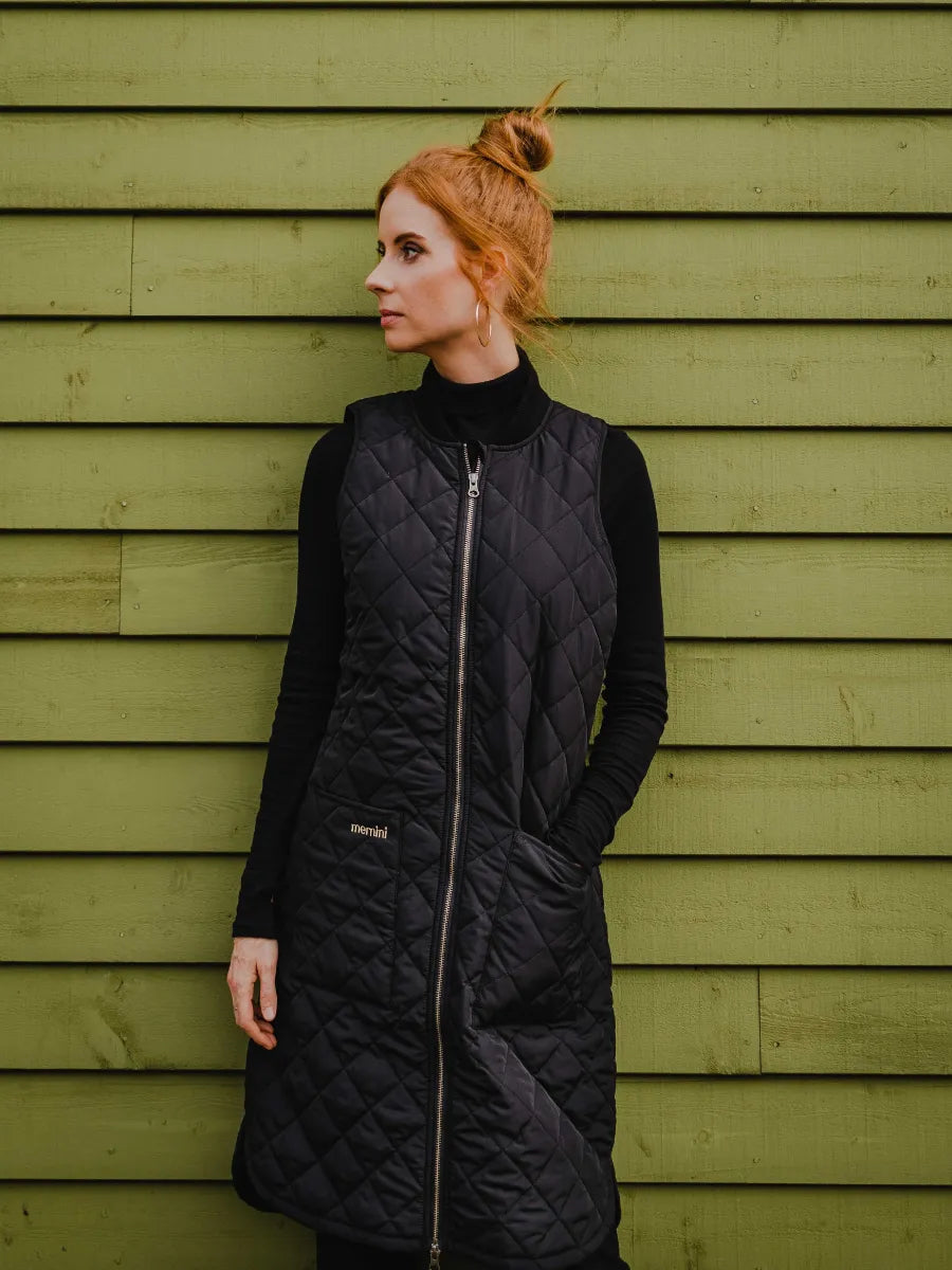 Person in a black quilted sleeveless long vest over a black top standing against an olive green wooden wall.