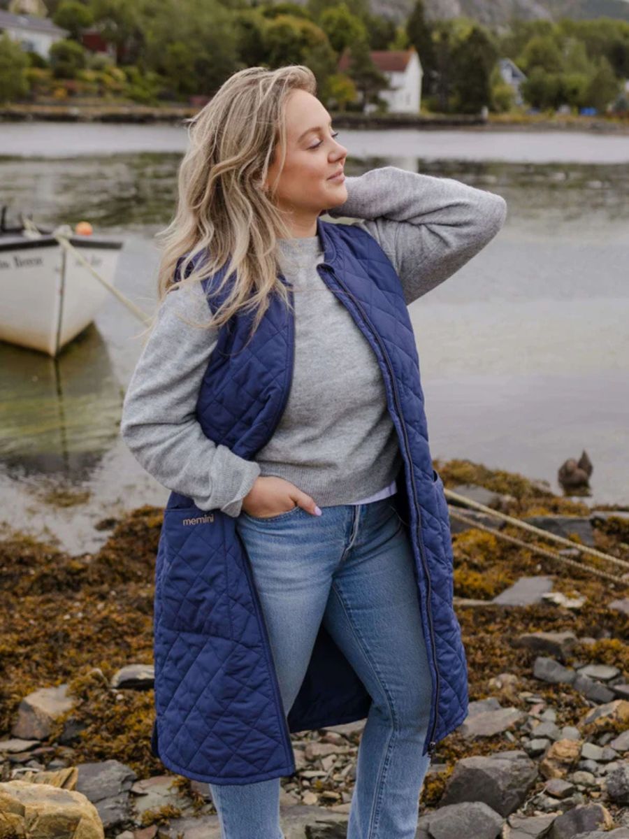 Person standing near water wearing a long quilted navy blue sleeveless vest over a gray sweater and jeans