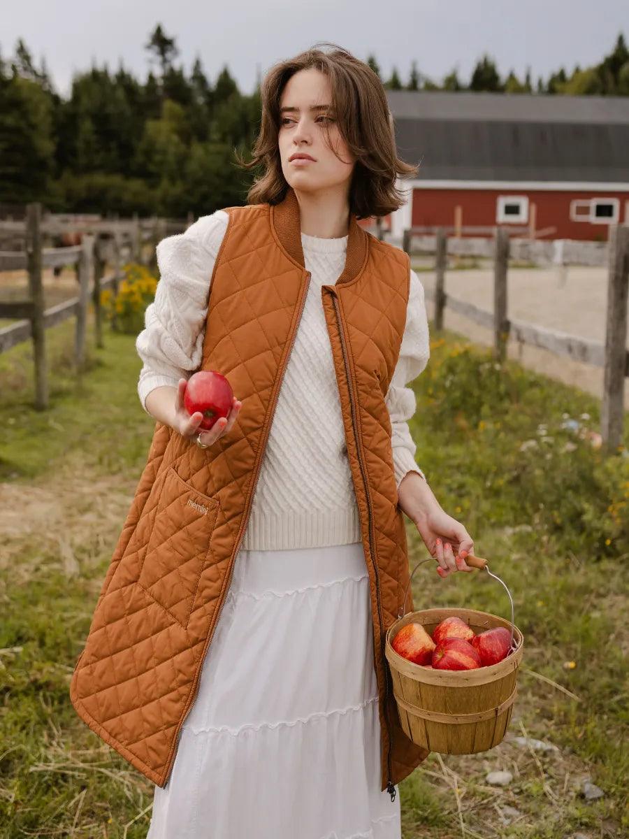 Person outdoors wearing a caramel quilted vest over a white sweater and skirt holding a basket of red apples