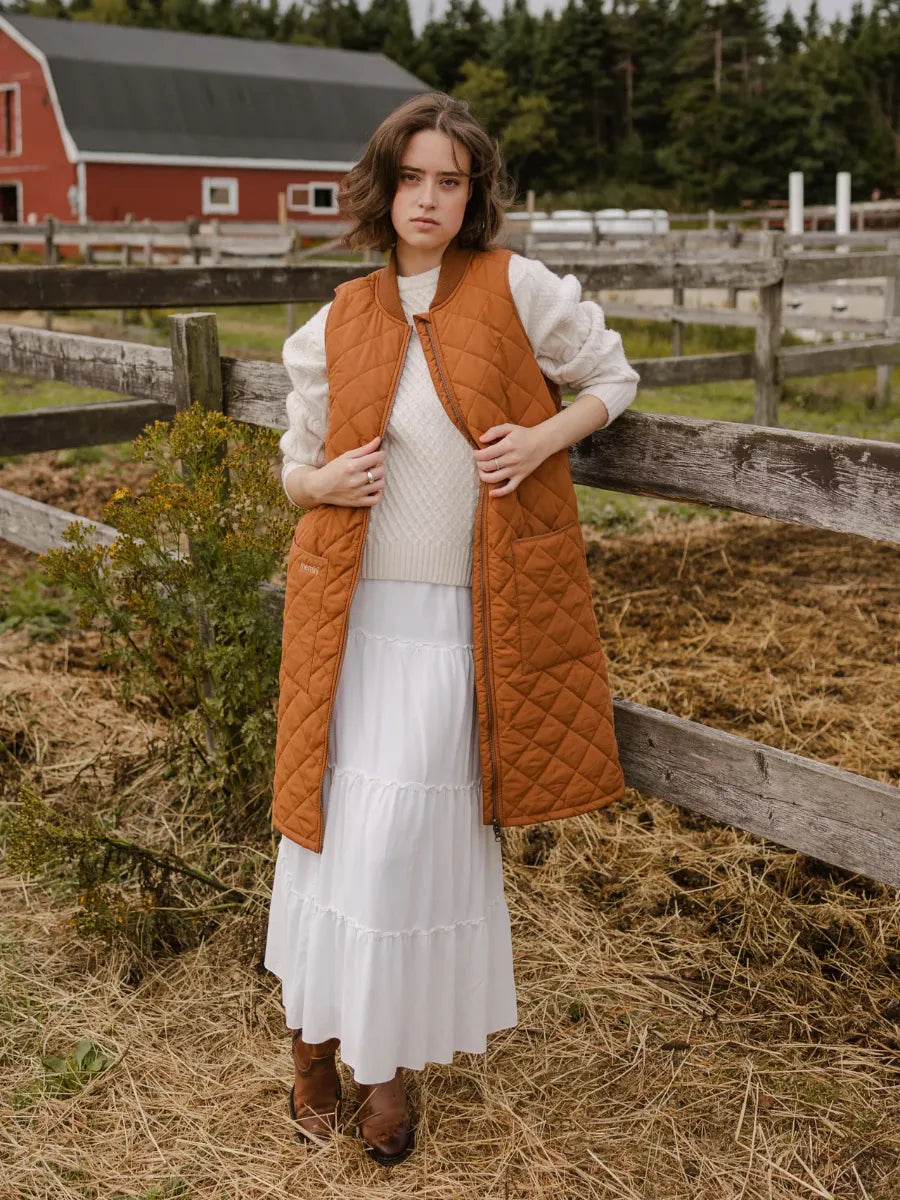 Person standing outdoors by wooden fence wearing a long caramel quilted vest over a white sweater and skirt