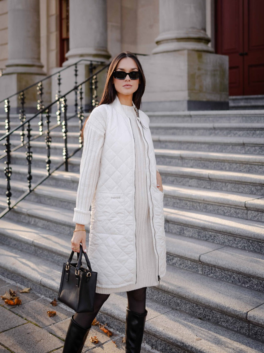 Person wearing a cream quilted longline vest over a cream sweater dress with black boots and sunglasses