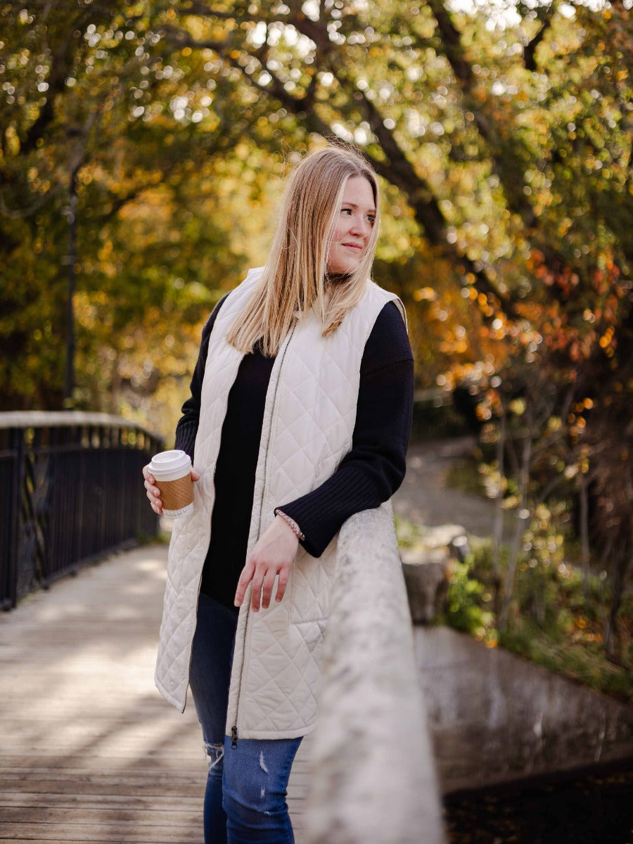 Person standing on a wooden bridge wearing a cream quilted vest and black sweater holding a coffee cup outdoors