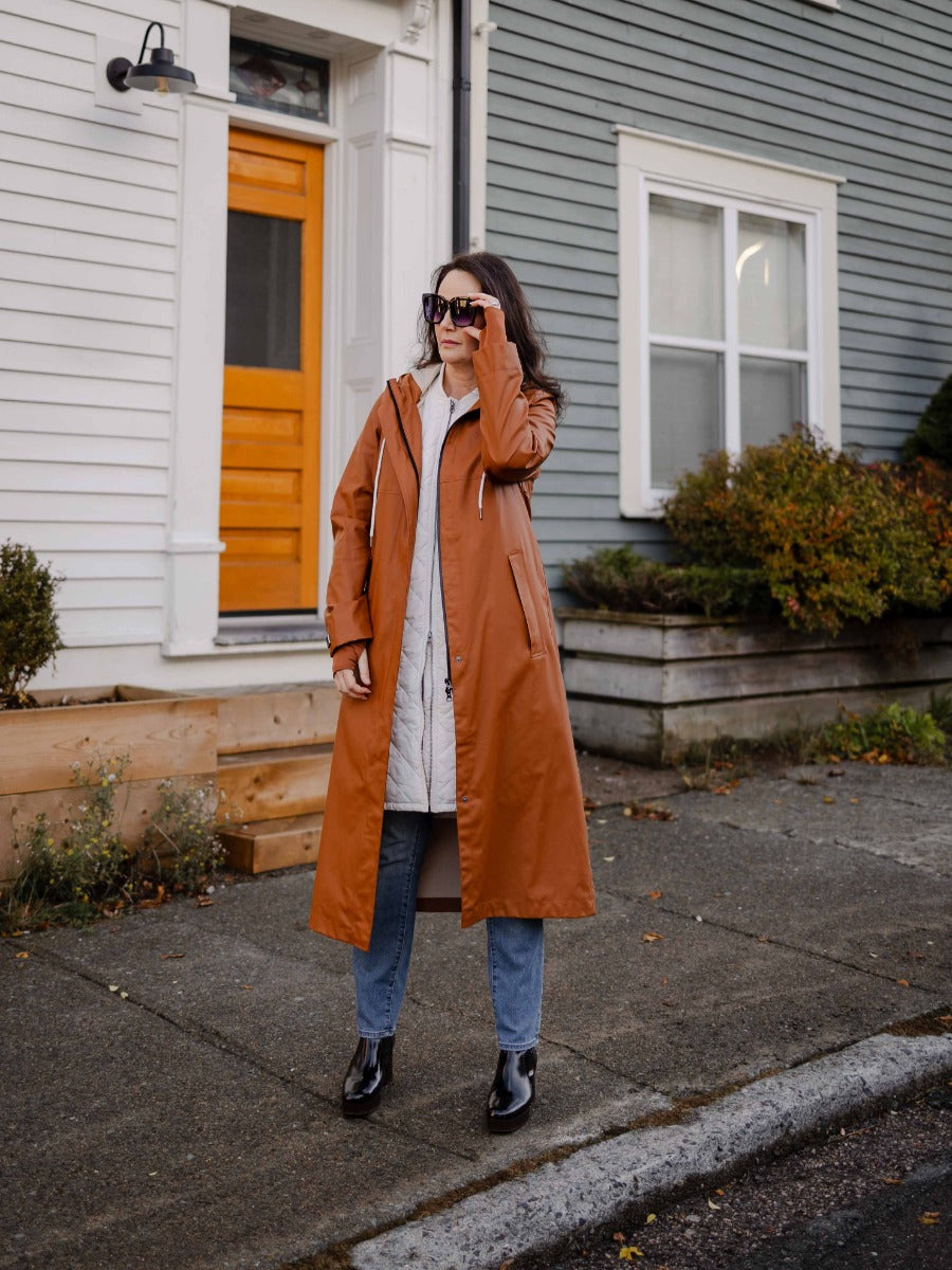 Person in long rust-colored coat and light quilted jacket stands on sidewalk in front of house with wooden door and gray siding