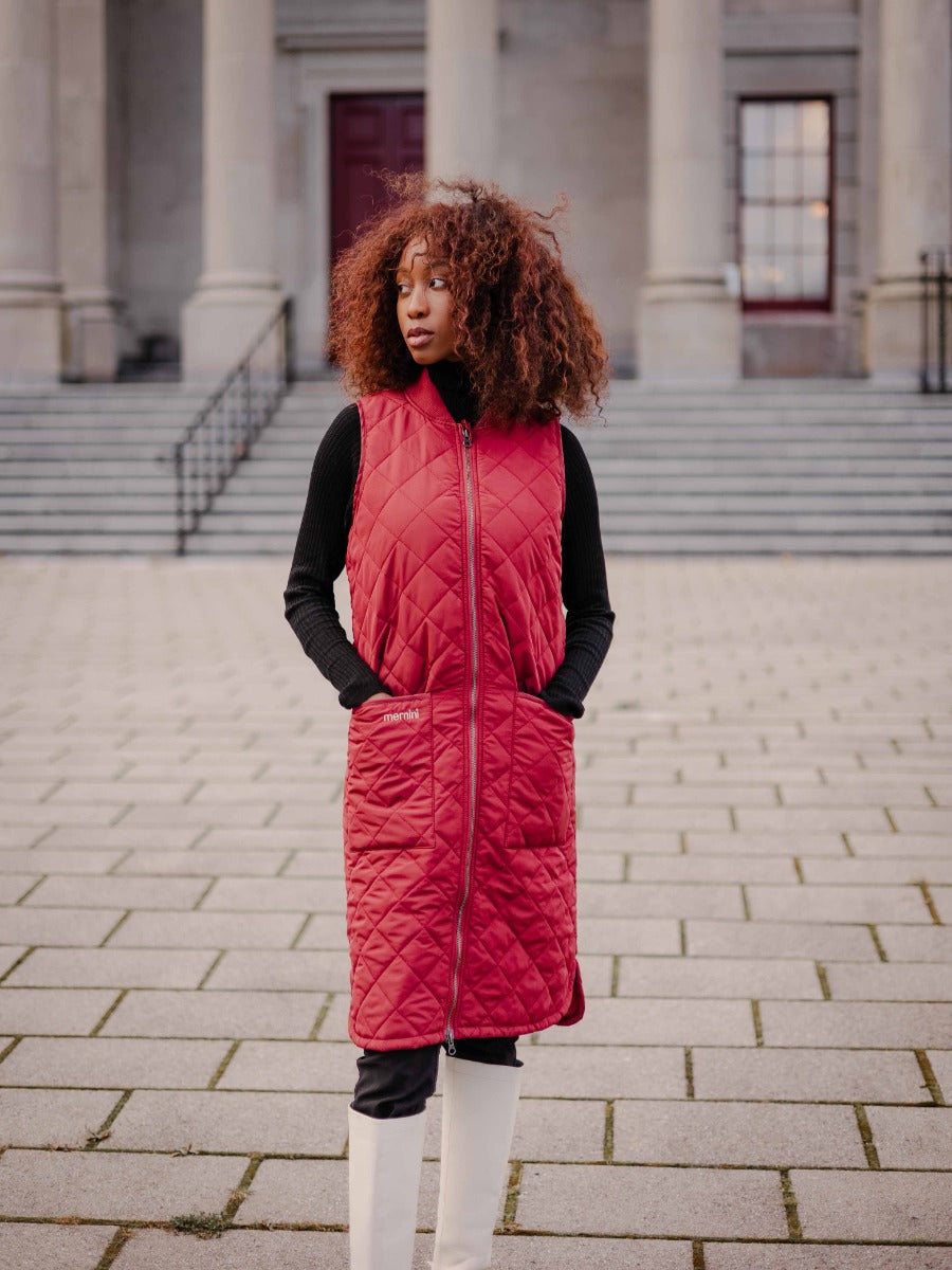 Model standing outdoors wearing a red Mernini Quilted Double Pocket Vest by MERNINI/ROCK RAIN INC over black top and white boots