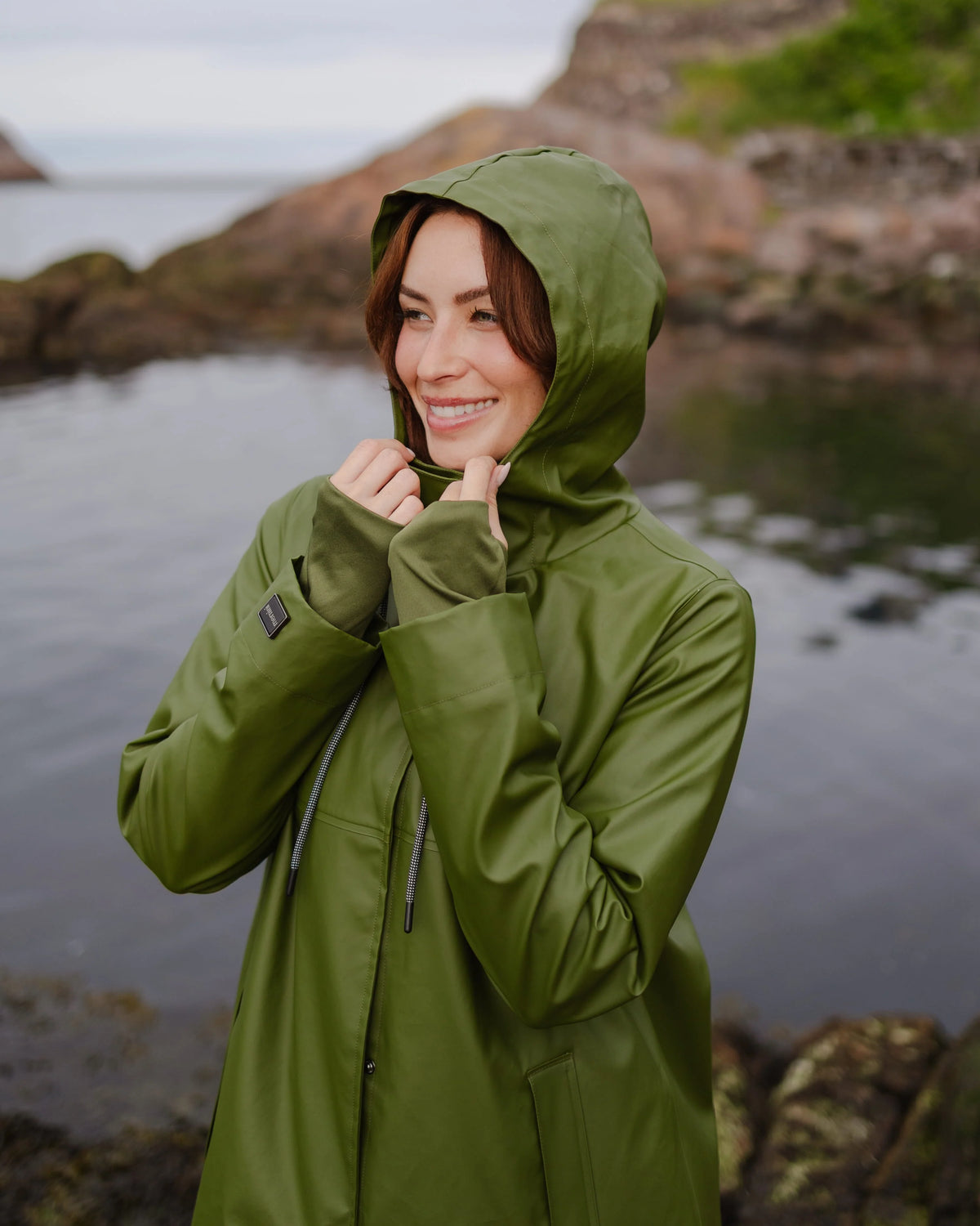 Smiling individual in a green hooded rain jacket beside a rocky coastal inlet