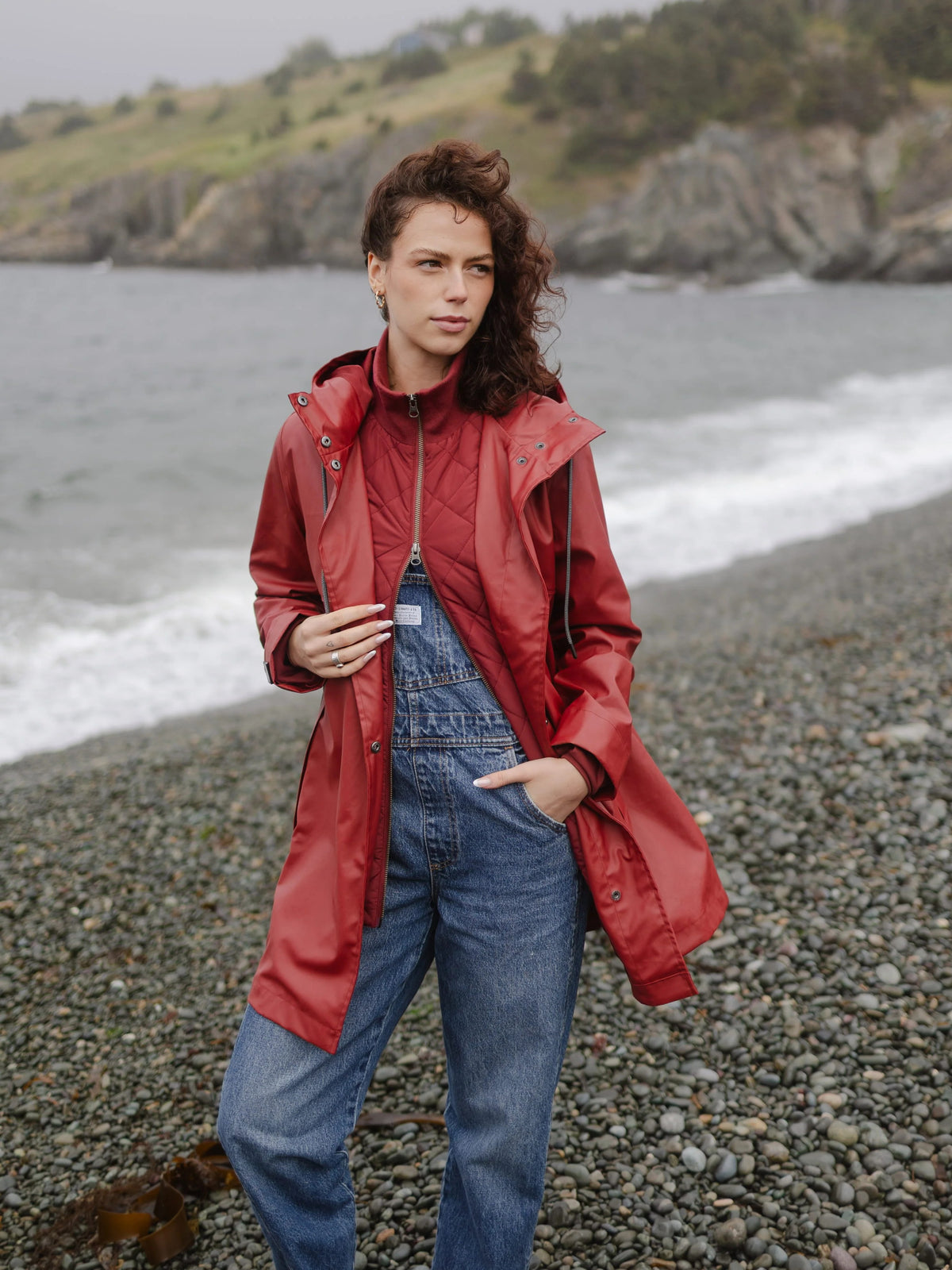 Model in a red raincoat and denim overalls standing on a rocky beach by the sea