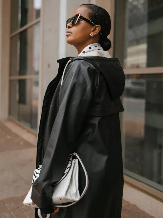 Profile view of a figure in a black hooded outerwear jacket, wearing large sunglasses and holding a white handbag.