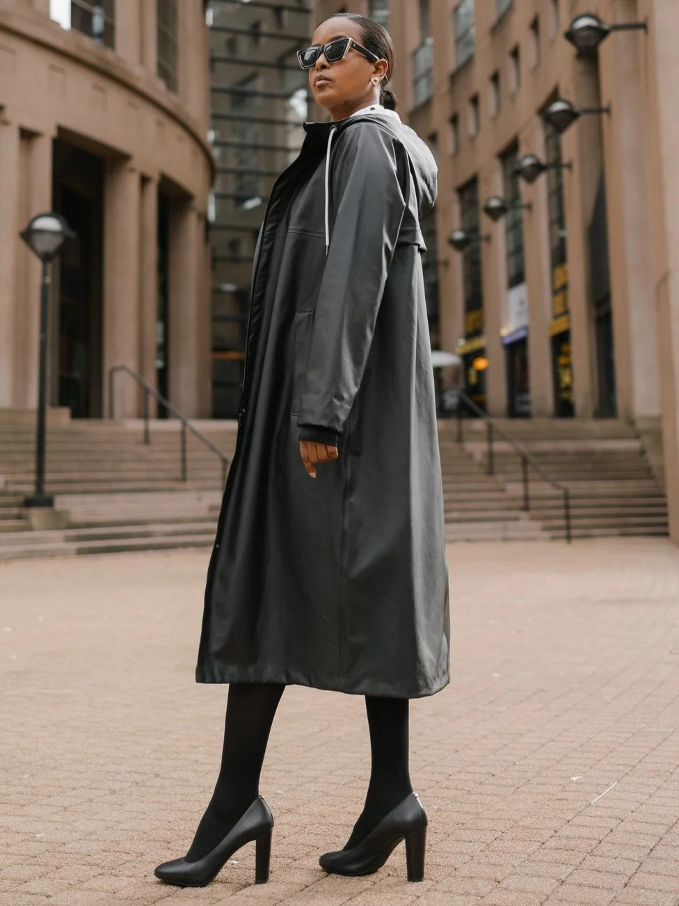 Individual in black long coat and high heels standing outdoors in an urban setting with steps and buildings in the background
