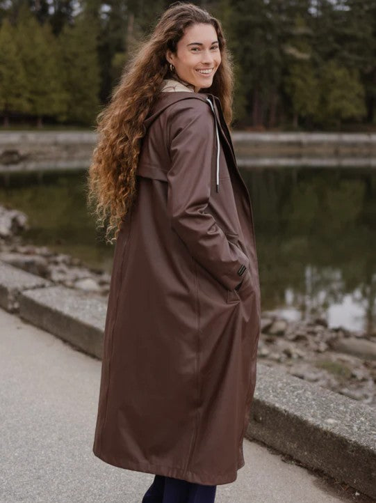 Person smiling outdoors wearing a long brown hooded coat with hands in pockets near water and trees
