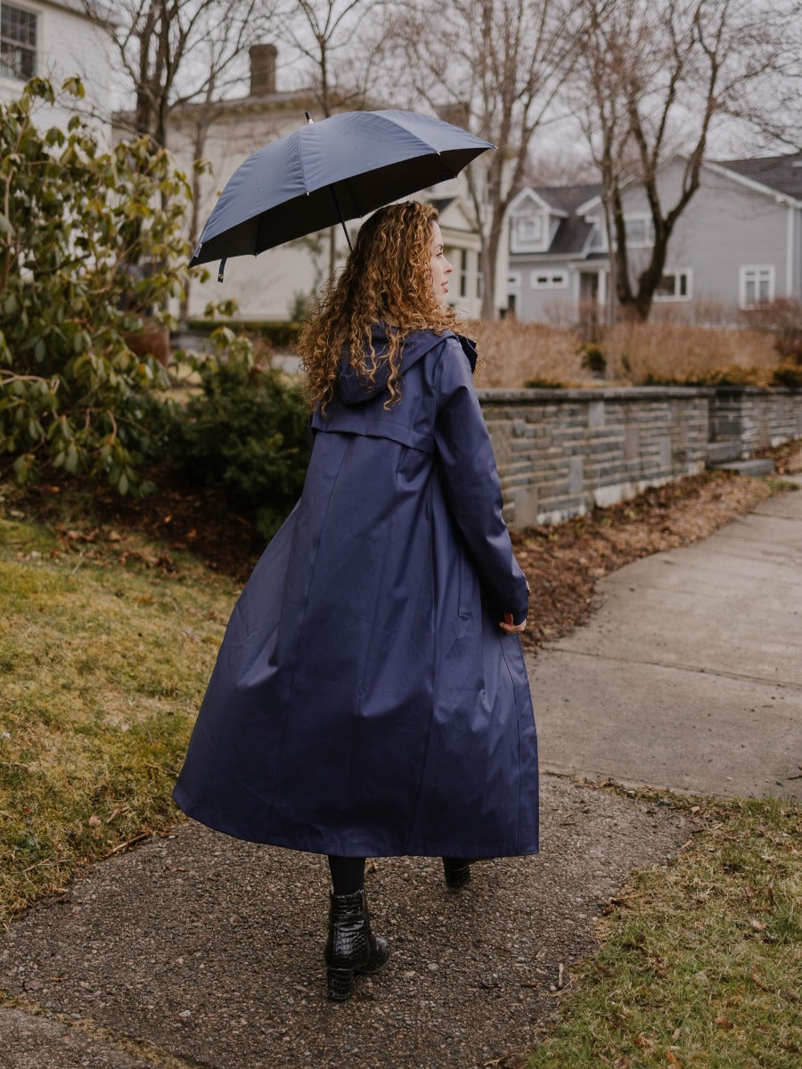 Person walking outdoors wearing a long navy coat and holding a matching umbrella on a cloudy day