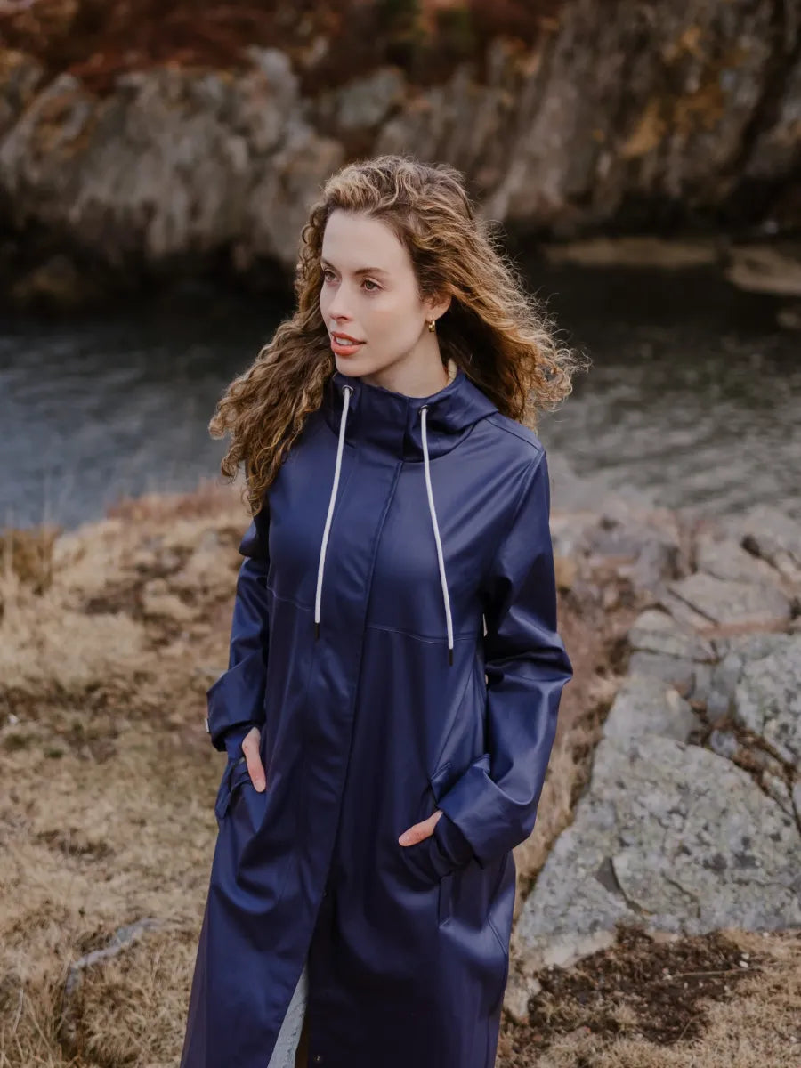 Person wearing a navy blue hooded jacket standing outdoors near rocky terrain and a body of water.