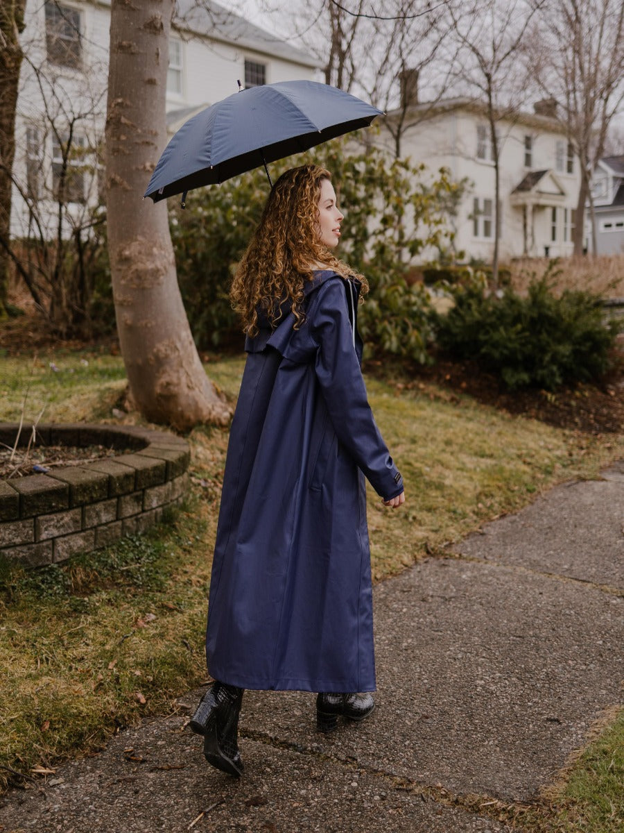 Person walking outdoors on a sidewalk holding a navy blue umbrella and wearing a long navy coat