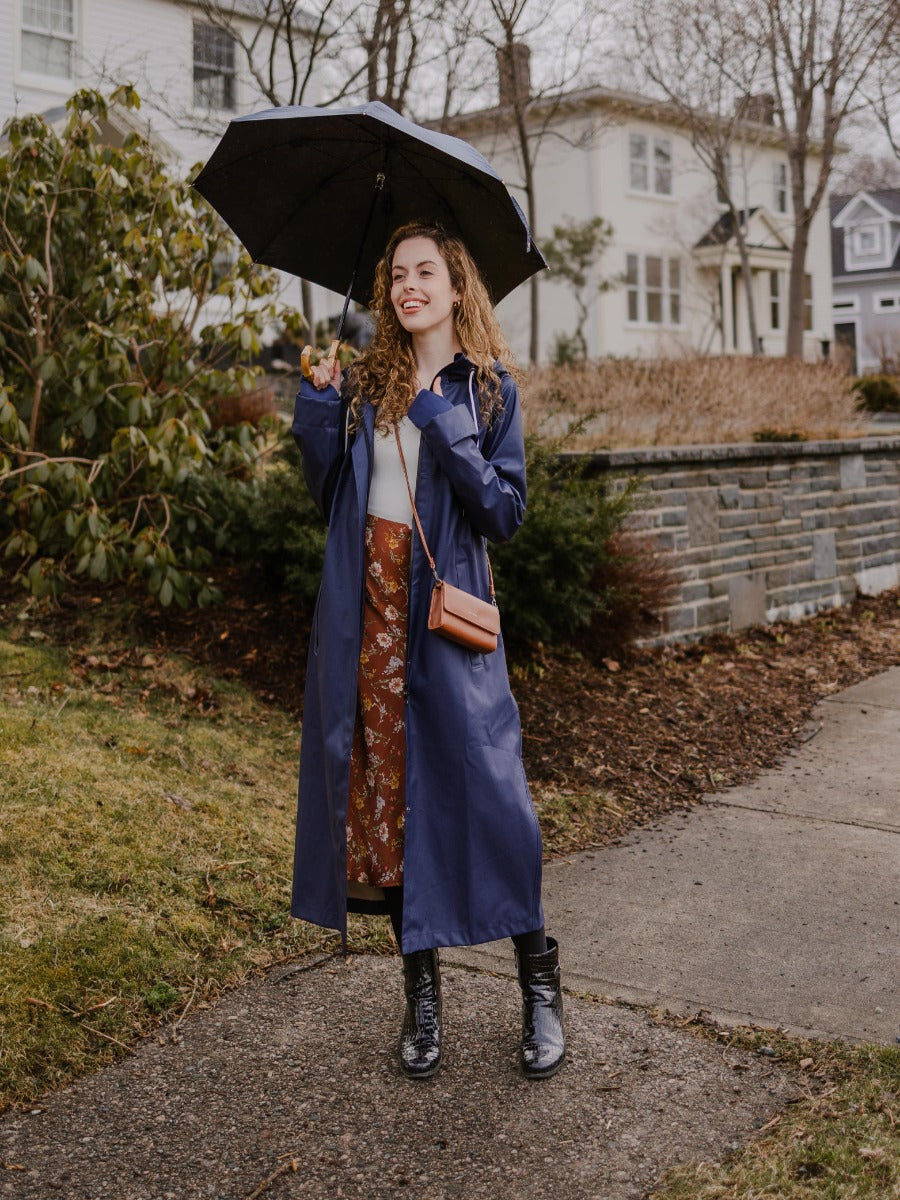 Person with long curly hair wearing a navy coat and skirt holding a black umbrella on a sidewalk in front of houses