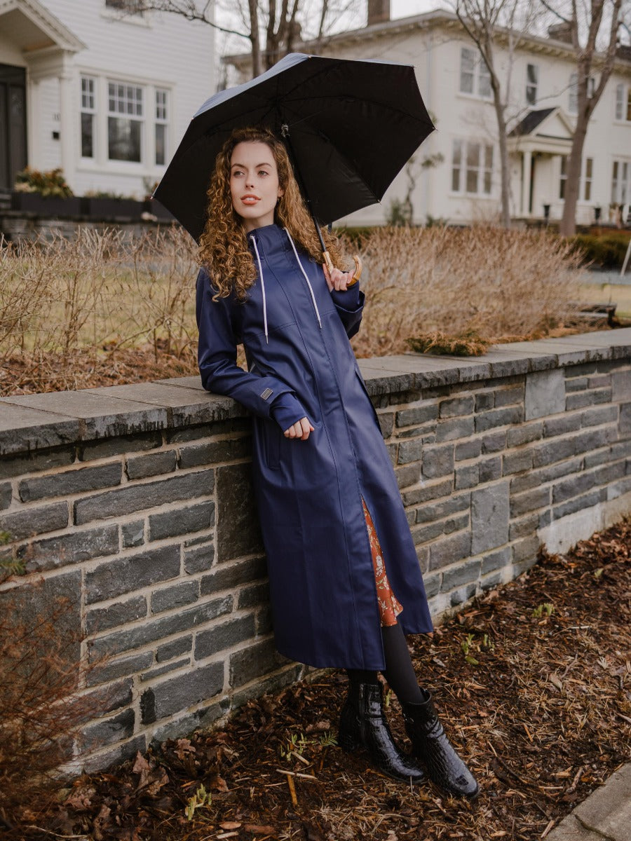 Model outdoors holding umbrella wearing navy blue Mernini 48” Length Waterproof Raincoat by MERNINI/ROCK RAIN INC.