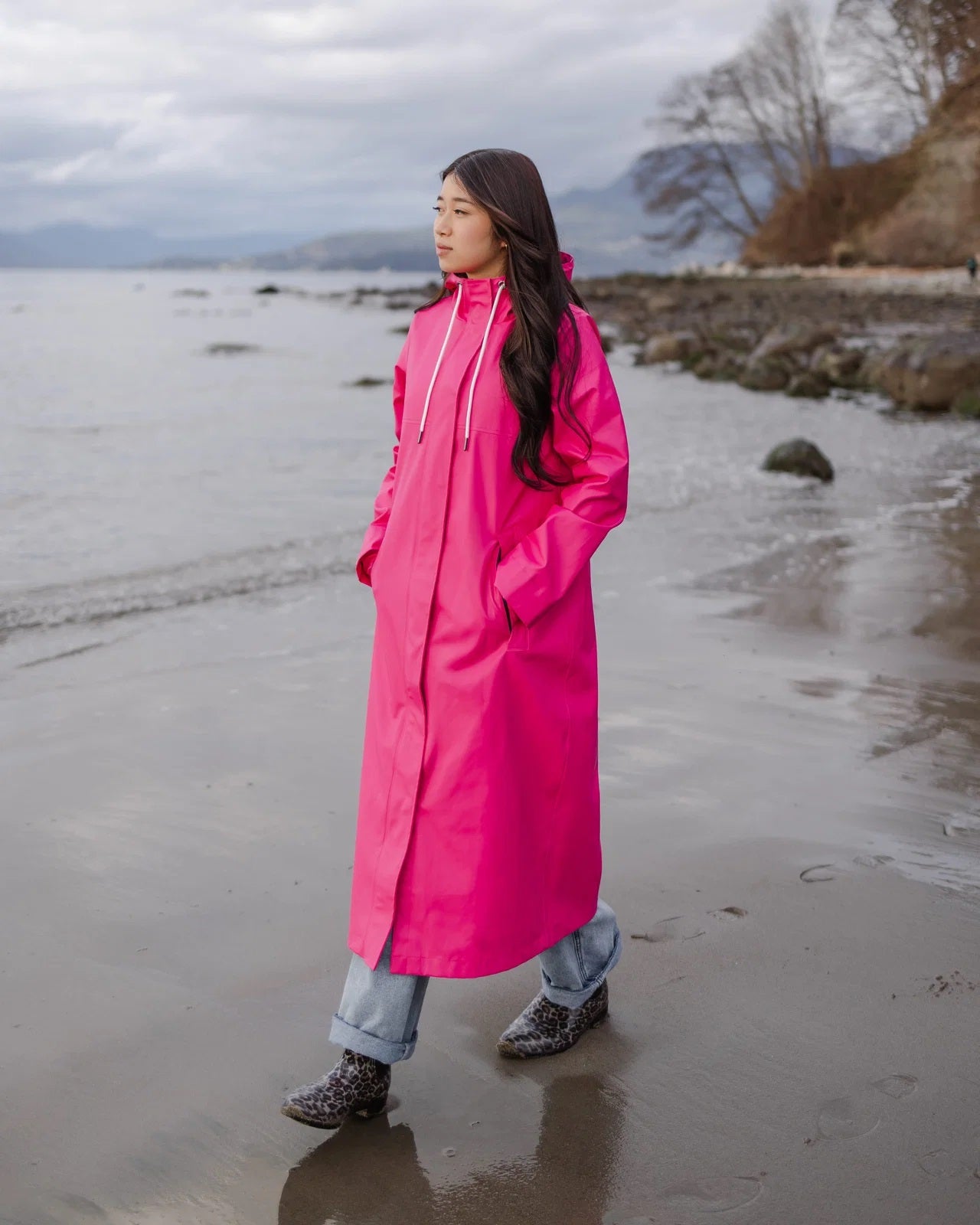 Long bright pink waterproof raincoat with hood worn outdoors on a beach with a cloudy sky background