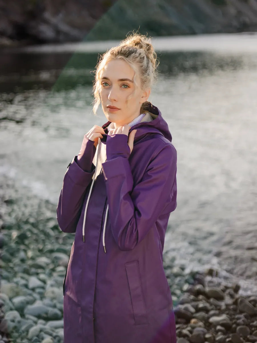 Person standing by a rocky riverbank wearing a long purple coat with a hood and white drawstrings.