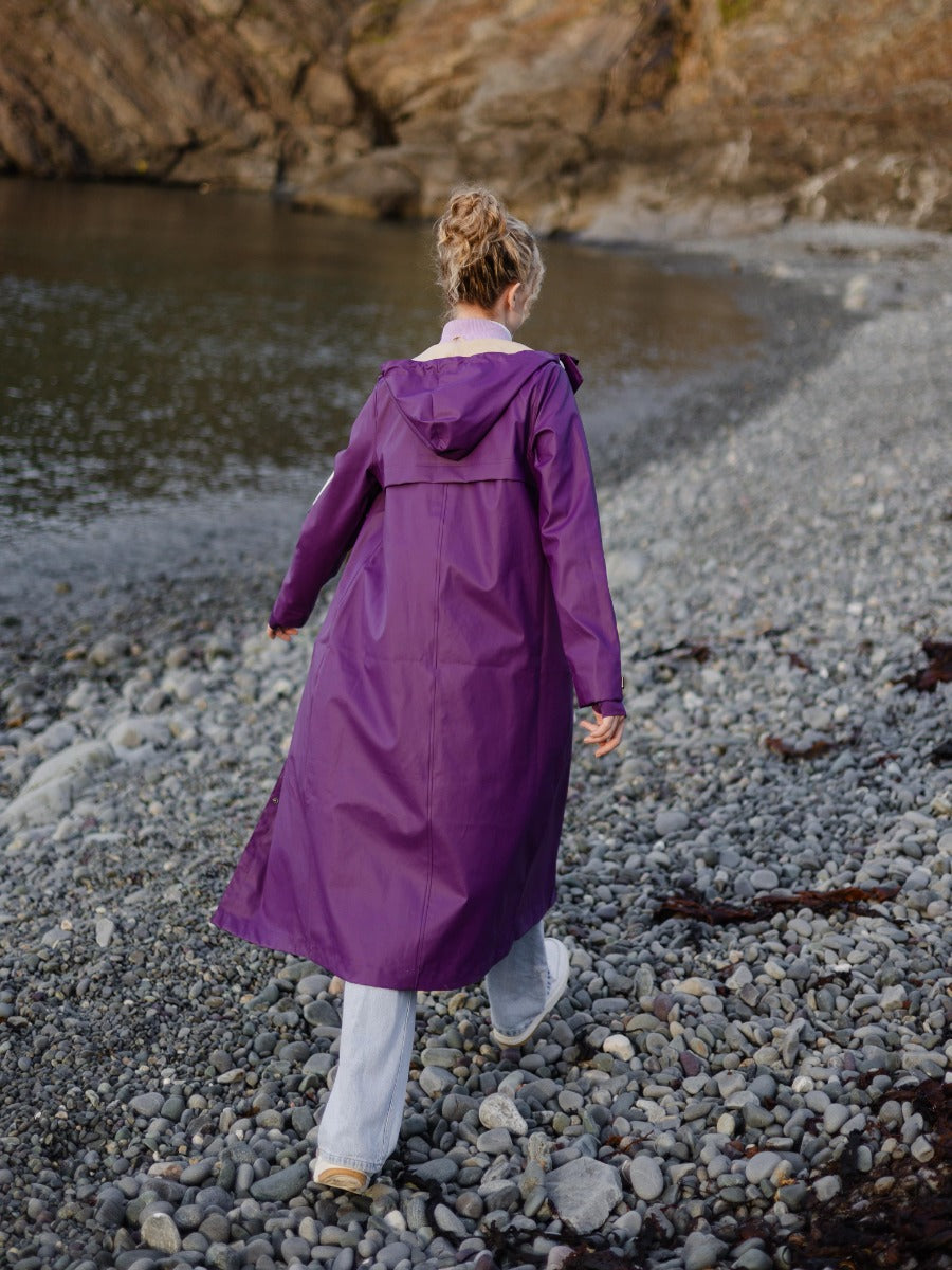 Person walking on rocky shorewearing a long purple raincoat with a hood and light-colored pants