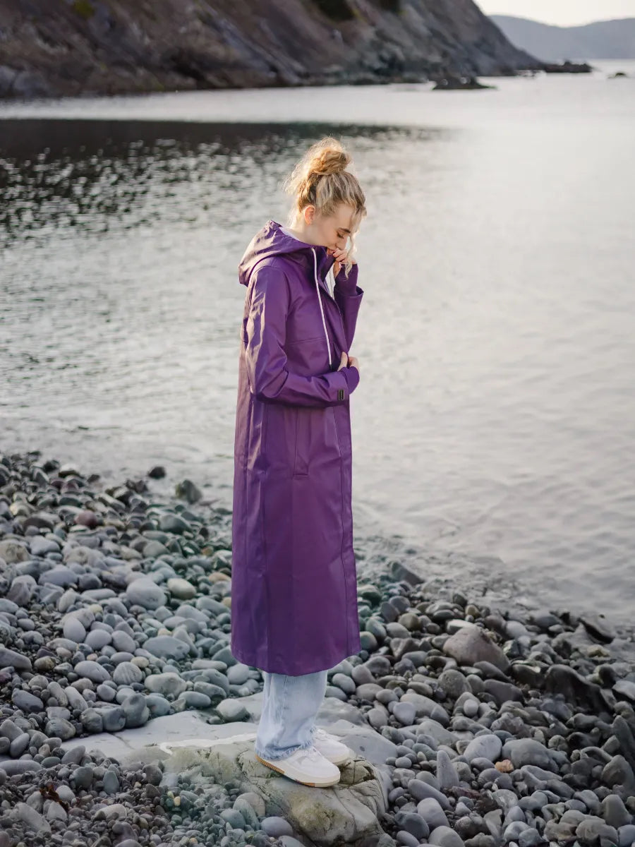 Person standing on rocky shore wearing a purple Mernini 48” Length Waterproof Raincoat by MERNINI/ROCK RAIN INC