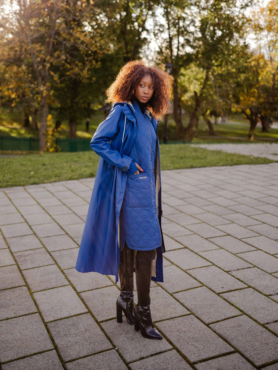 Person wearing a long blue quilted coat layered over a trench coat with black heeled boots outdoors in autumn.