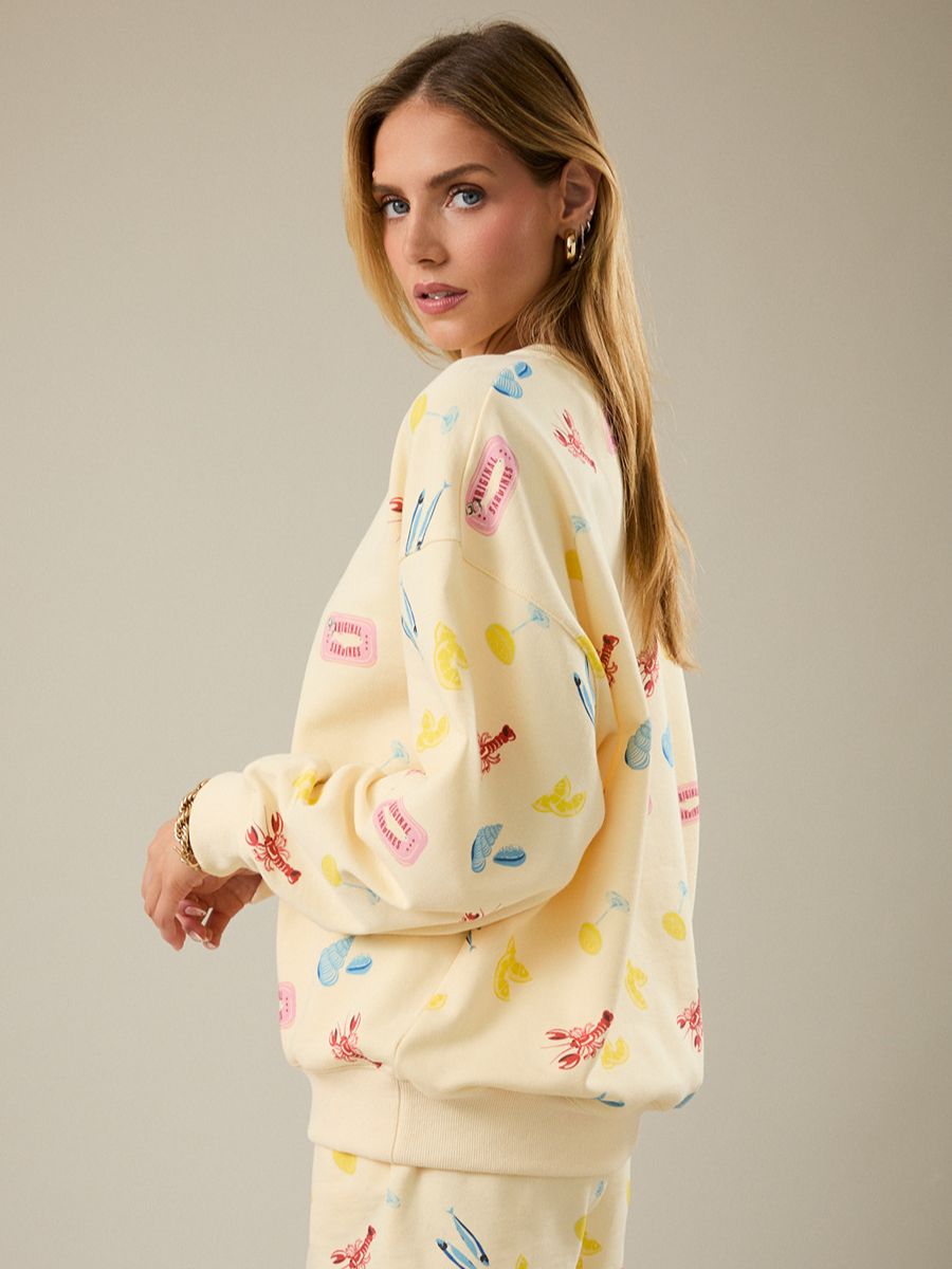 Light yellow sweatshirt with colorful playful prints of lobsters, shoes, and other small objects on a neutral background.