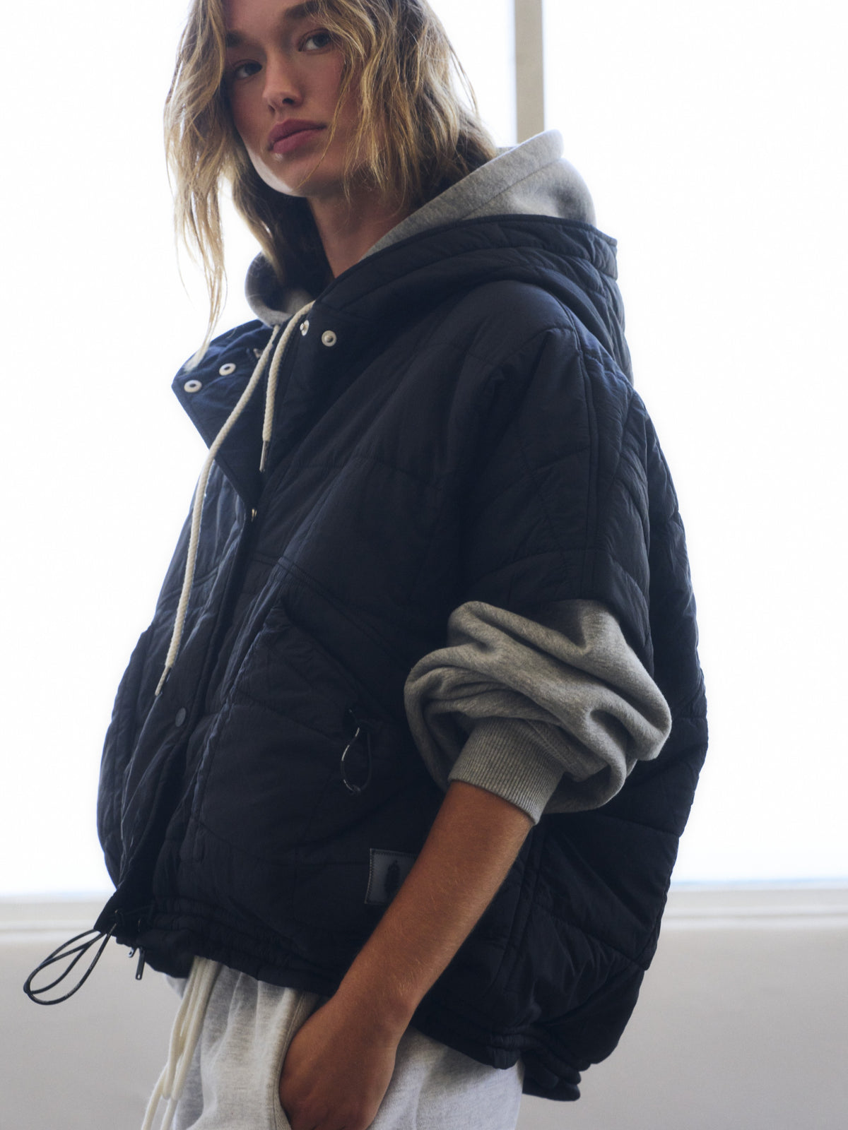 Blond figure in a black quilted hooded jacket layered over a gray sweatshirt by a window