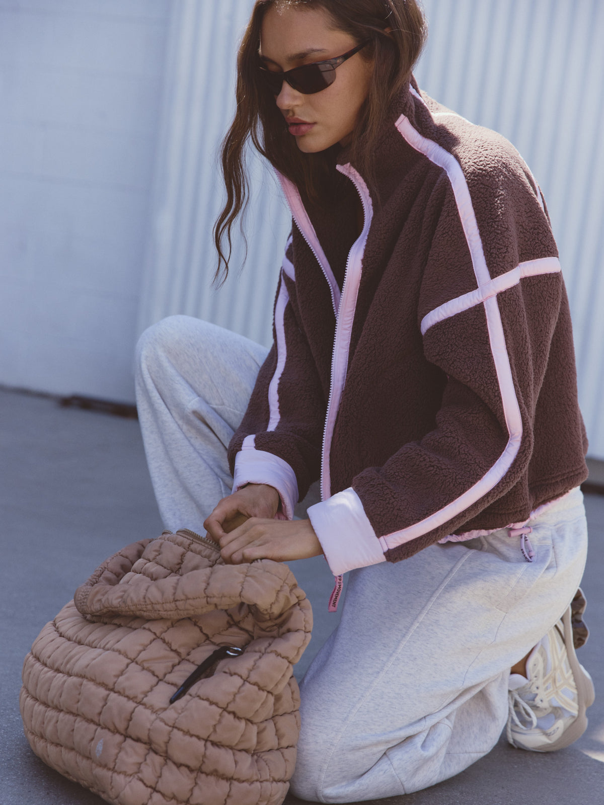 Brown fleece jacket with pink trim, light jeans, sunglasses, and a quilted brown bag outdoors