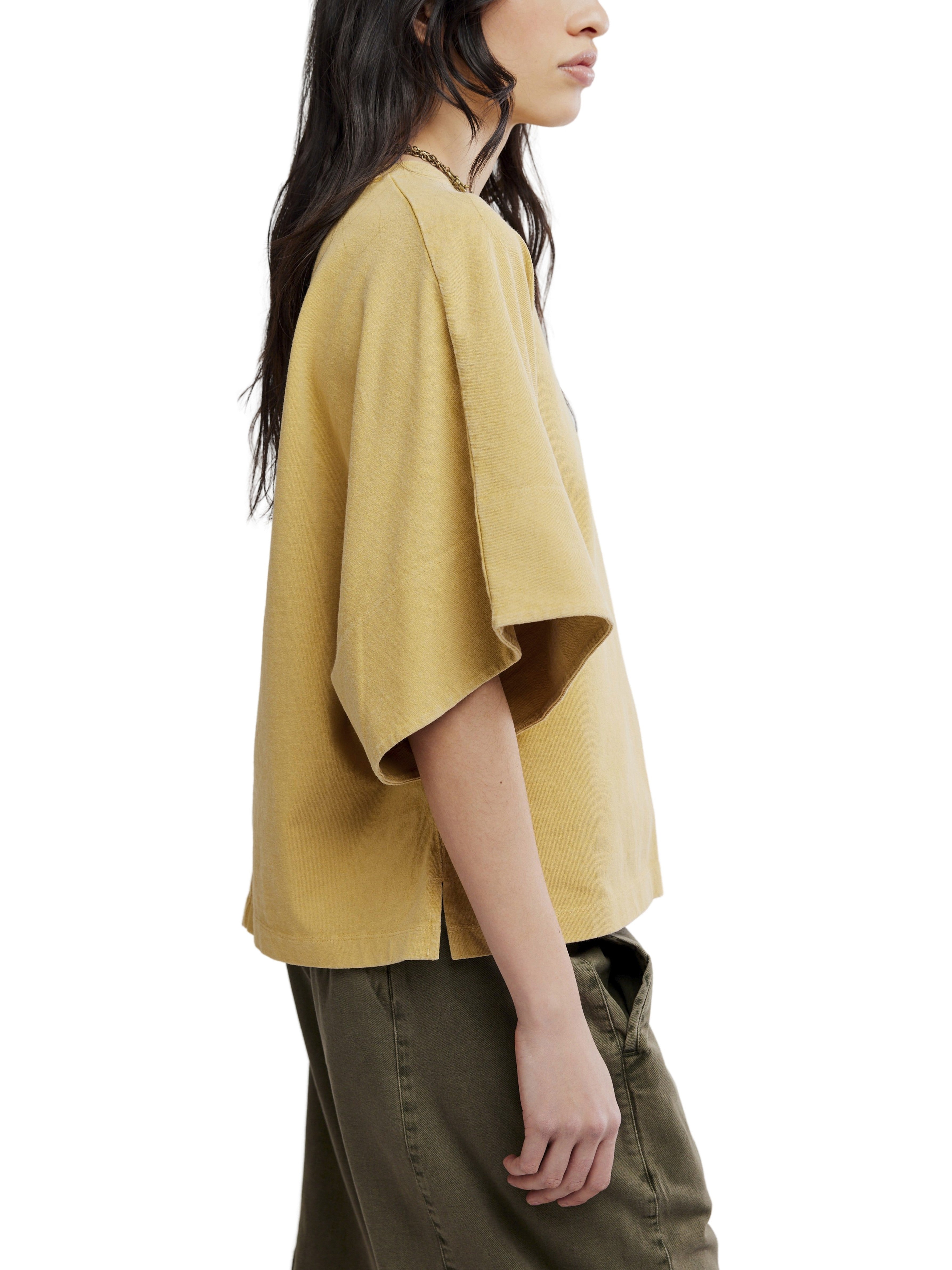 Side view of a person wearing a loose mustard yellow short-sleeve top and olive green pants against a white background.