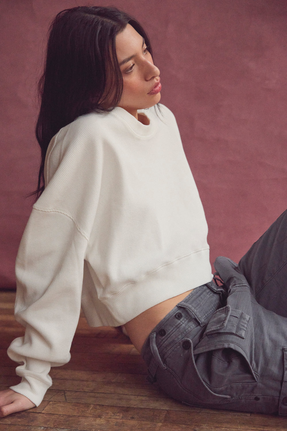 Woman in a cream sweater and gray jeans seated against a muted pink backdrop