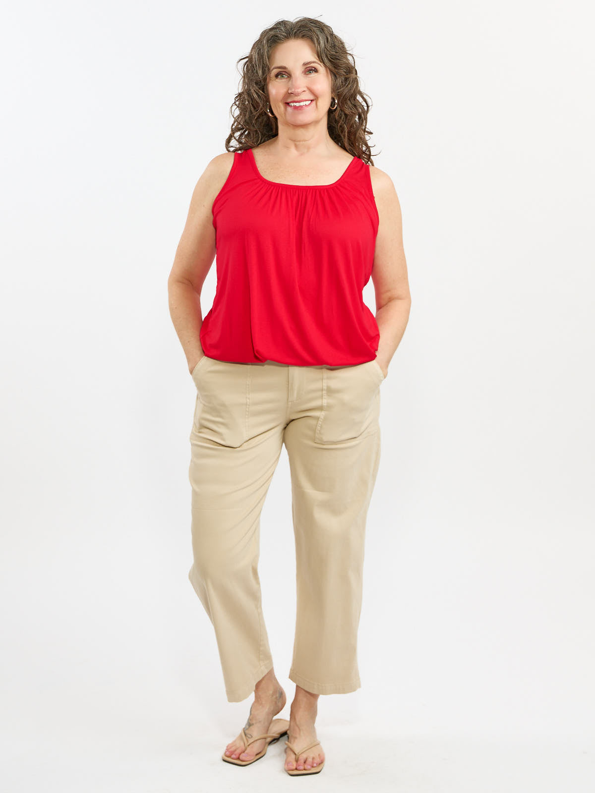 Curly-haired individual in a sleeveless red top, beige pants with pockets, and sandals standing against white background.