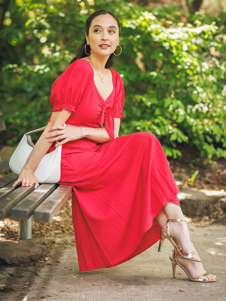 Elegant red Papillon Bunny Tie Dress paired with gold sandals and white handbag, seated on a park bench outdoors.