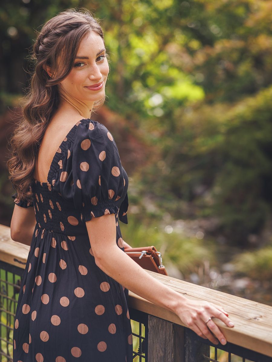 Papillon Polka Dot Bunny Tie Midi Dress in black with taupe dots, puff sleeves, and shirred back detail outdoors.