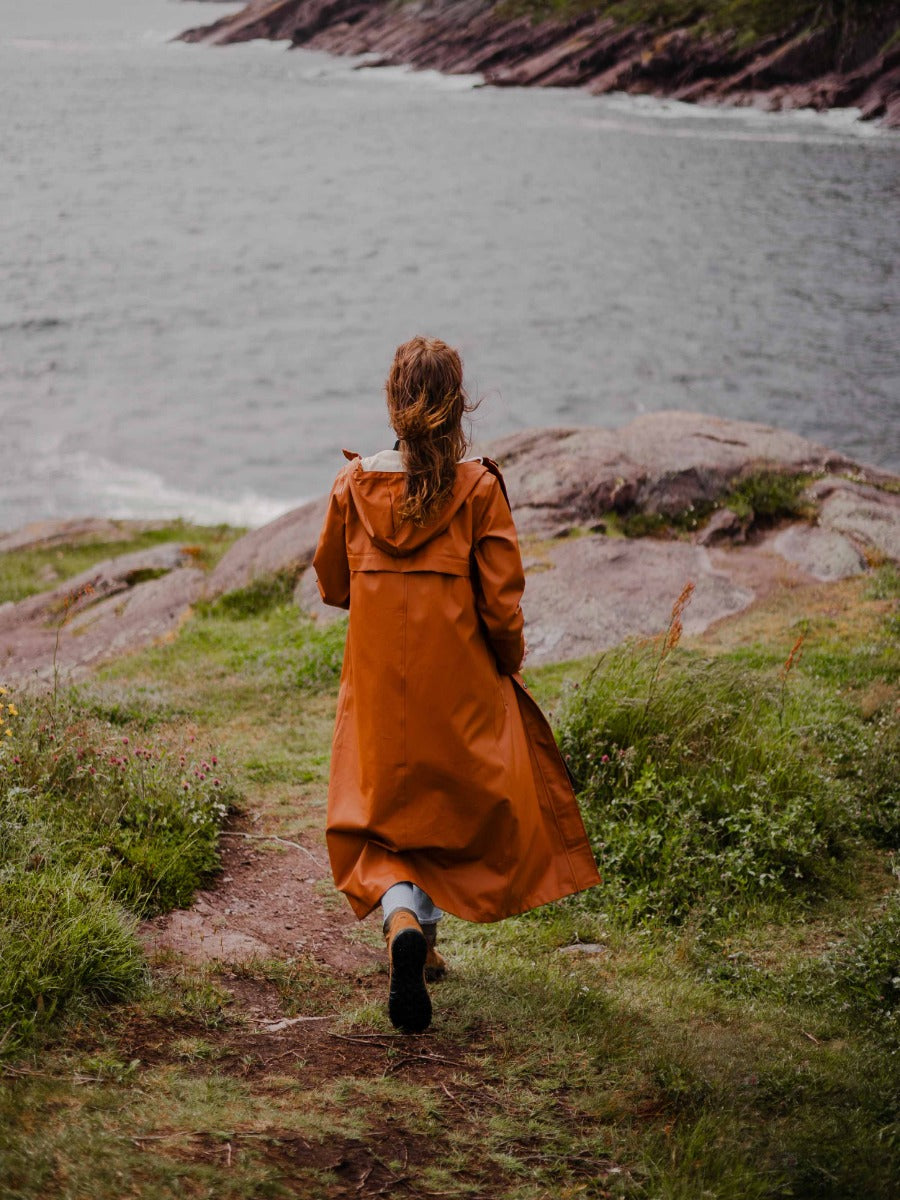 Person wearing a long rust-colored coat walking on a grassy path near rocky shores by the water