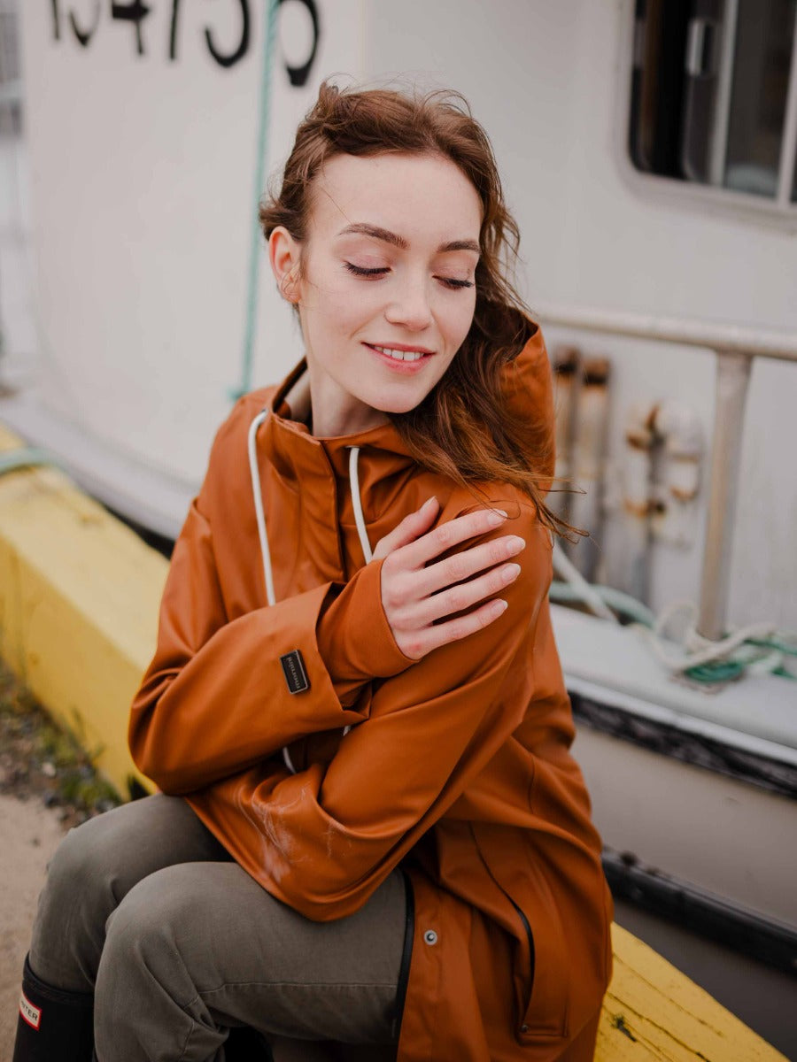 Person sitting outdoors wearing a caramel-colored waterproof jacket with white drawstrings by a boat dock