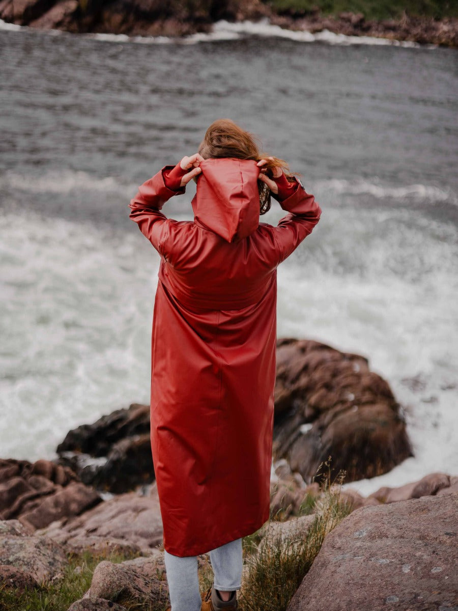 Person adjusting the hood of a long red raincoat standing on rocky coastline near choppy water