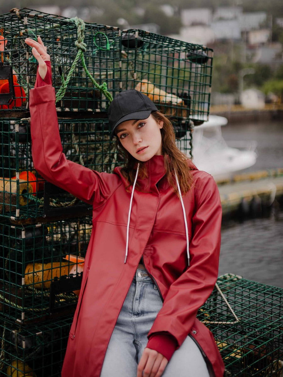 Person in a red coat and black cap sitting by stacked fishing traps on a dock near water and boats.