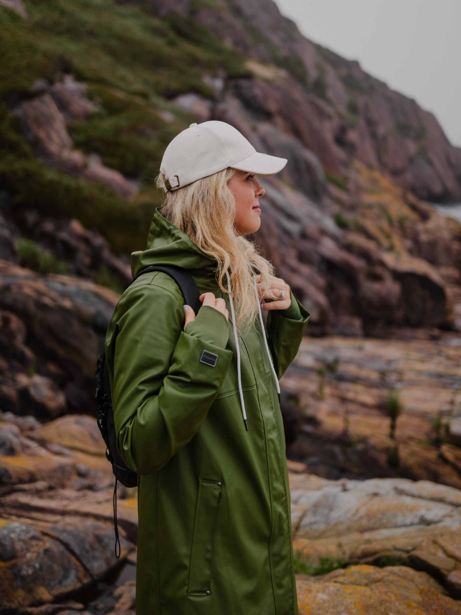 Person in olive green raincoat and white cap standing on rocky terrain with grassy hillside in the background