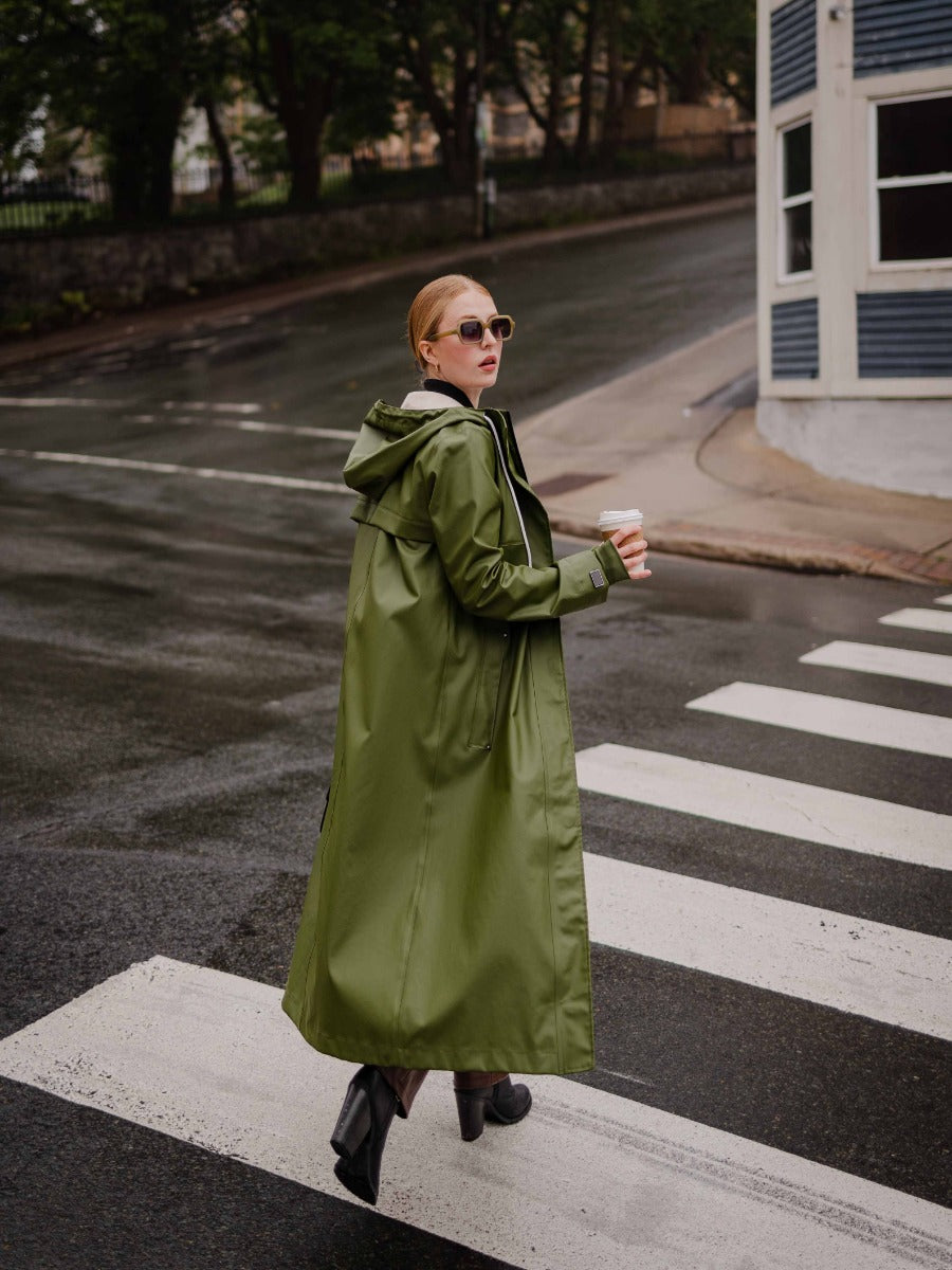 Person crossing street wearing long olive green raincoat and sunglasses, holding a coffee cup on wet pavement.