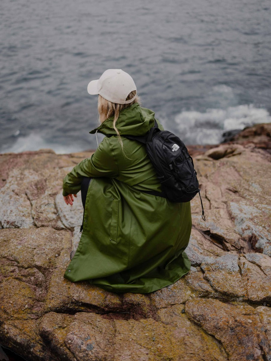 Person sitting on rocky shore wearing olive green coat, beige cap, and black backpack facing the ocean
