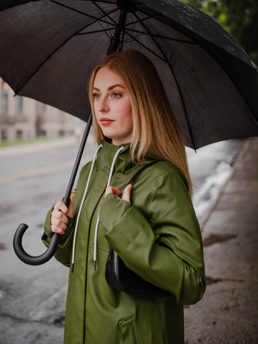 Person holding umbrella wearing MERNINI/ROCK RAIN INC Mernini Petite 42” Length Waterproof Raincoat in olive green on rainy street